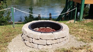 A lakeside fire pit area with a chain-link fence, surrounded by a grassy area and a wooden deck. - White River Bank Cabin!Eagle Watching, World Class Trout Fishing Off  The Dock,  (Flippin)