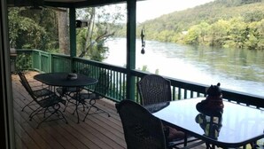 A covered outdoor deck with a table and chairs, overlooking a river and forested hills. - White River Bank Cabin!Eagle Watching, World Class Trout Fishing Off  The Dock,  (Flippin)