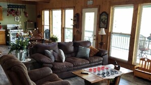 A cozy cabin interior with a brown leather sofa, a coffee table with a checkered board game, and large windows offering views of the outdoors. - White River Bank Cabin!Eagle Watching, World Class Trout Fishing Off  The Dock,  (Flippin)