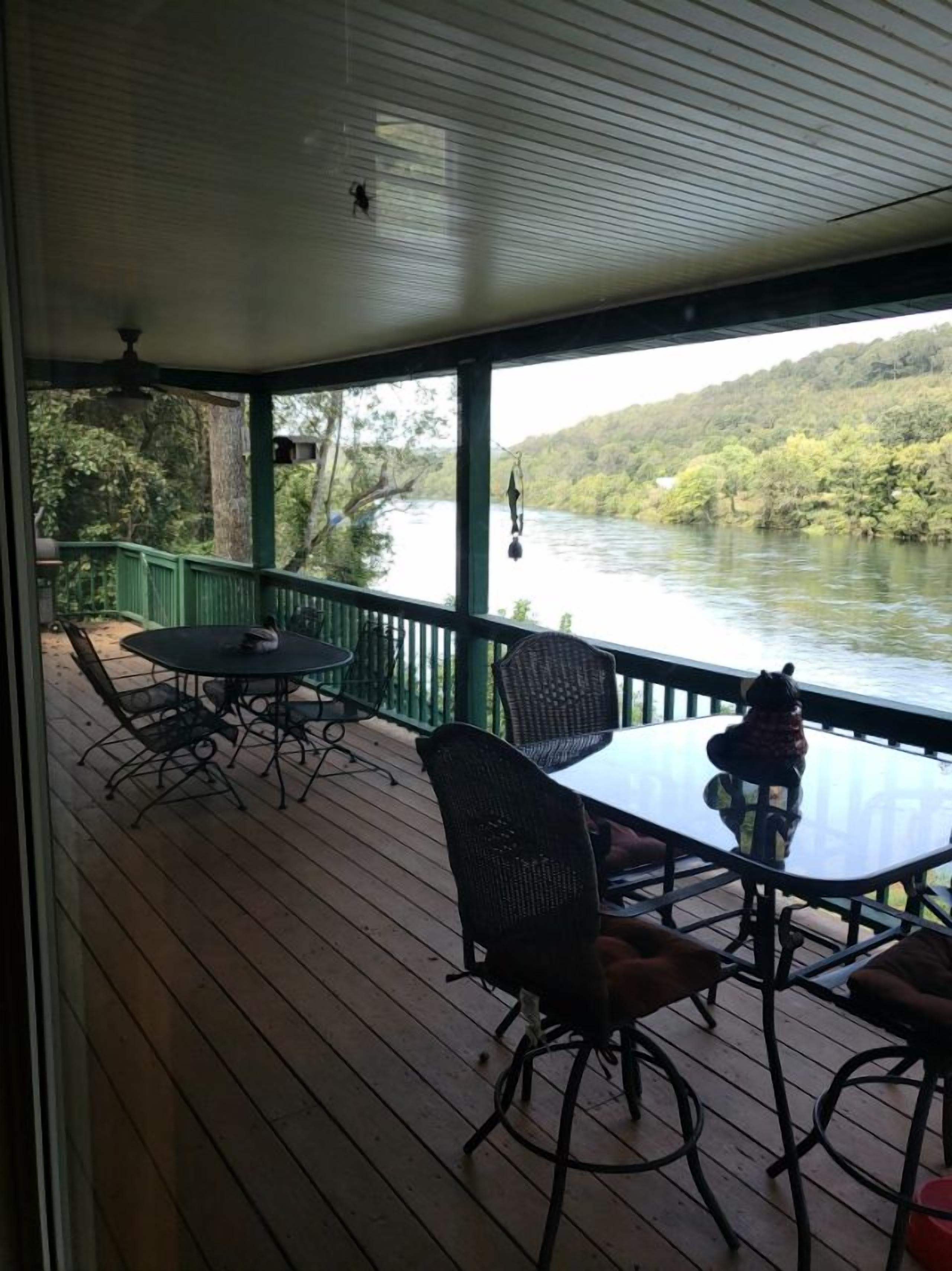 Une terrasse extérieure couverte, dotée d’une table et de chaises, qui offre une vue sur une rivière et des collines boisées.