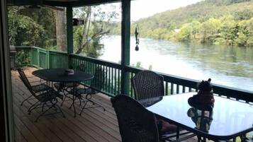 Une terrasse extérieure couverte, dotée d’une table et de chaises, qui offre une vue sur une rivière et des collines boisées.