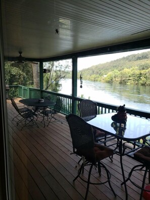 A covered outdoor deck with a table and chairs, overlooking a river and forested hills. - White River Bank Cabin!Eagle Watching, World Class Trout Fishing Off The Dock, (Flippin)