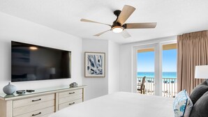 Een slaapkamer met een bed, een plafondventilator, een televisie, een dressoir, een raam met uitzicht op het strand en een schilderij aan de muur.