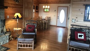 L’intérieur d’un chalet rustique, avec un plancher en bois, un coin repas avec des chaises, une lampe et une cheminée agrémentée d’un coussin à motifs d’ours.
