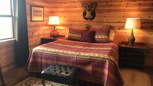 Une pièce chaleureuse avec un mur en bois, un lit recouvert d’une couverture colorée, deux tables de chevet avec des lampes, une tête de hibou décorative et un tapis à motifs.