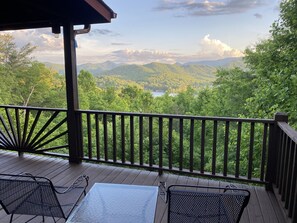 A wooden deck with metal chairs and a table, overlooking a lush green landscape with mountains and a lake. - Last Min Disc Pictures Don’t Do View Justice Hot tub. Pool Table. Easy to get to (Hiawassee)