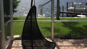 Una silla colgante negra en un espacio exterior techado, con vista a un cuerpo de agua y un puente al fondo.