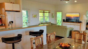 A modern kitchen with a refrigerator, dining table, and chairs. There is a television on a stand and a window with a view of greenery.