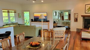 A living room with a sofa, a dining table with a fruit bowl, a television, a fireplace, and a view of the outdoors.