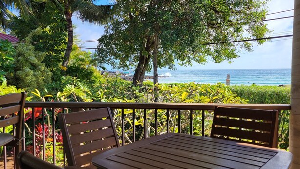 A wooden deck with two chairs and a table, overlooking a beach with clear blue water and palm trees.