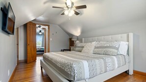 A bedroom with a large bed, a ceiling fan, a TV mounted on the wall, and a door leading to another room.