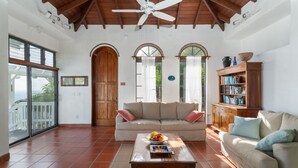 Un vaste espace de vie doté d’un plafond haut, de poutres en bois, d’un ventilateur de plafond, d’un canapé, d’une table basse ornée de fruits et d’une bibliothèque.