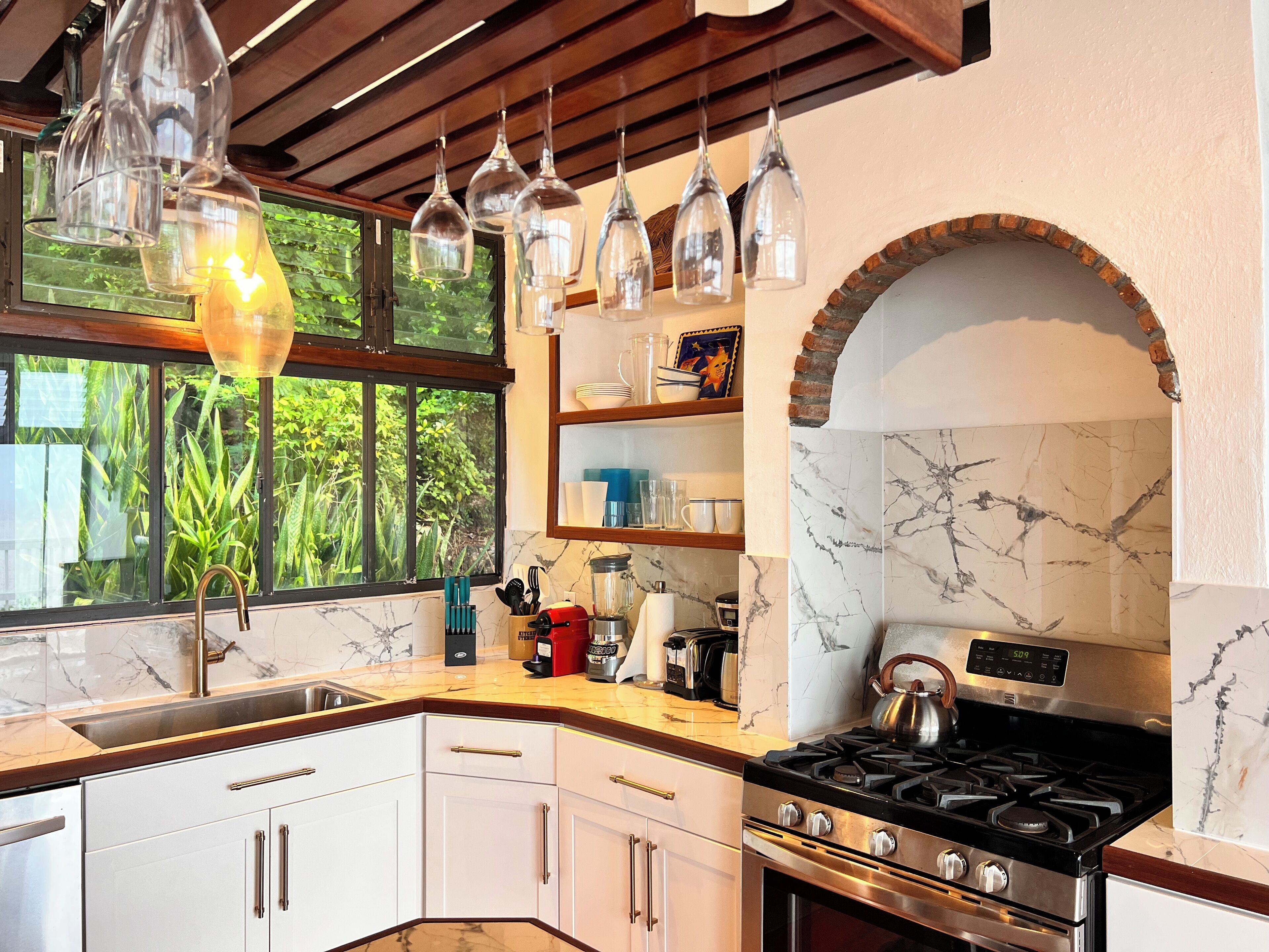 A modern kitchen with a stove, oven, and various appliances. There is a window with a view of greenery outside.