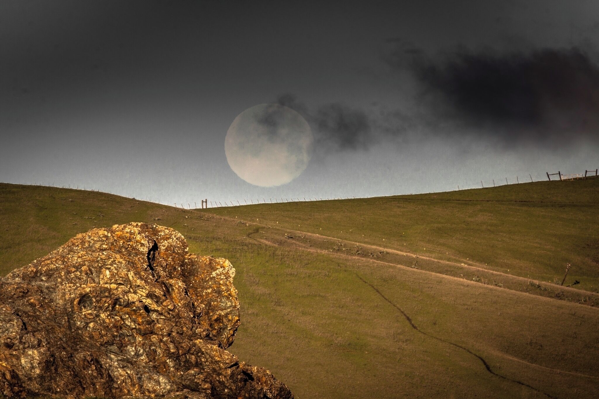 A rocky outcrop with a person standing on it, a fence in the background, and a large moon in the sky.