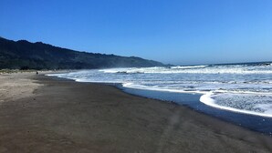 Une plage avec des vagues, un ciel dégagé et une colline boisée en arrière-plan.