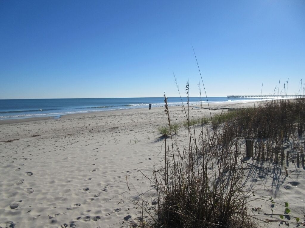 Une plage de sable avec des empreintes de pas, une végétation clairsemée et une jetée visible au loin.