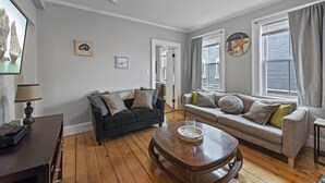 A living room with two sofas, a coffee table, and a TV. There are pictures on the wall and a window with curtains.