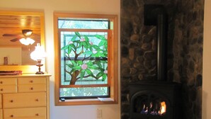 Een kamer met een houten dressoir, een glas-in-loodraam met een boom en een stenen openhaard met zichtbaar vuur.