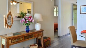 A room with a wooden console table, a vase with flowers, a mirror, and a lamp. There is a chair and a door leading to another room.