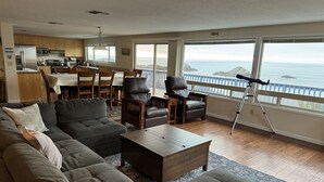 A spacious living room with a large sectional sofa, a coffee table, and two leather chairs. A dining area with wooden chairs and a table is visible in the background. The room features large windows with a view of the ocean. - Welcome to the Harris Beach House! ON the beach with private beach access. (Brookings)