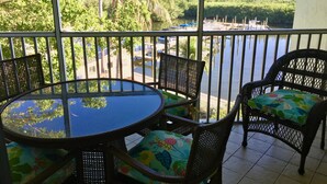 A balcony with a round table and wicker chairs, overlooking a body of water and greenery.