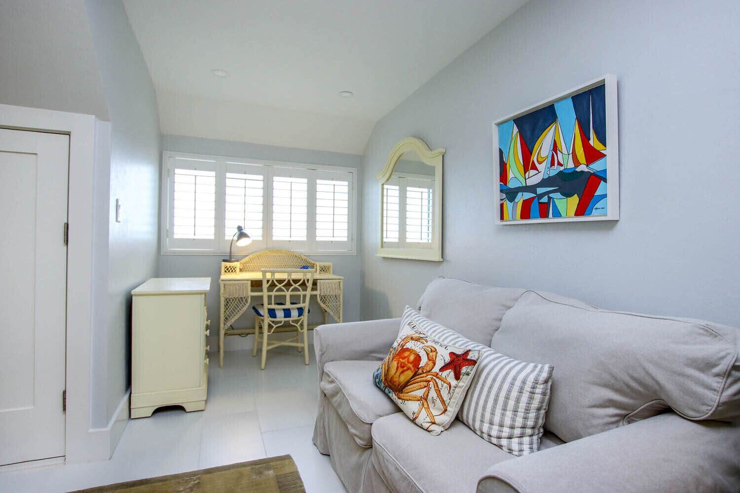A room with a white desk, a white chair, a grey sofa, a mirror, and a colorful painting on the wall.