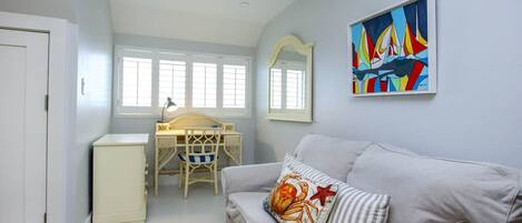 A room with a white desk, a white chair, a grey sofa, a mirror, and a colorful painting on the wall.