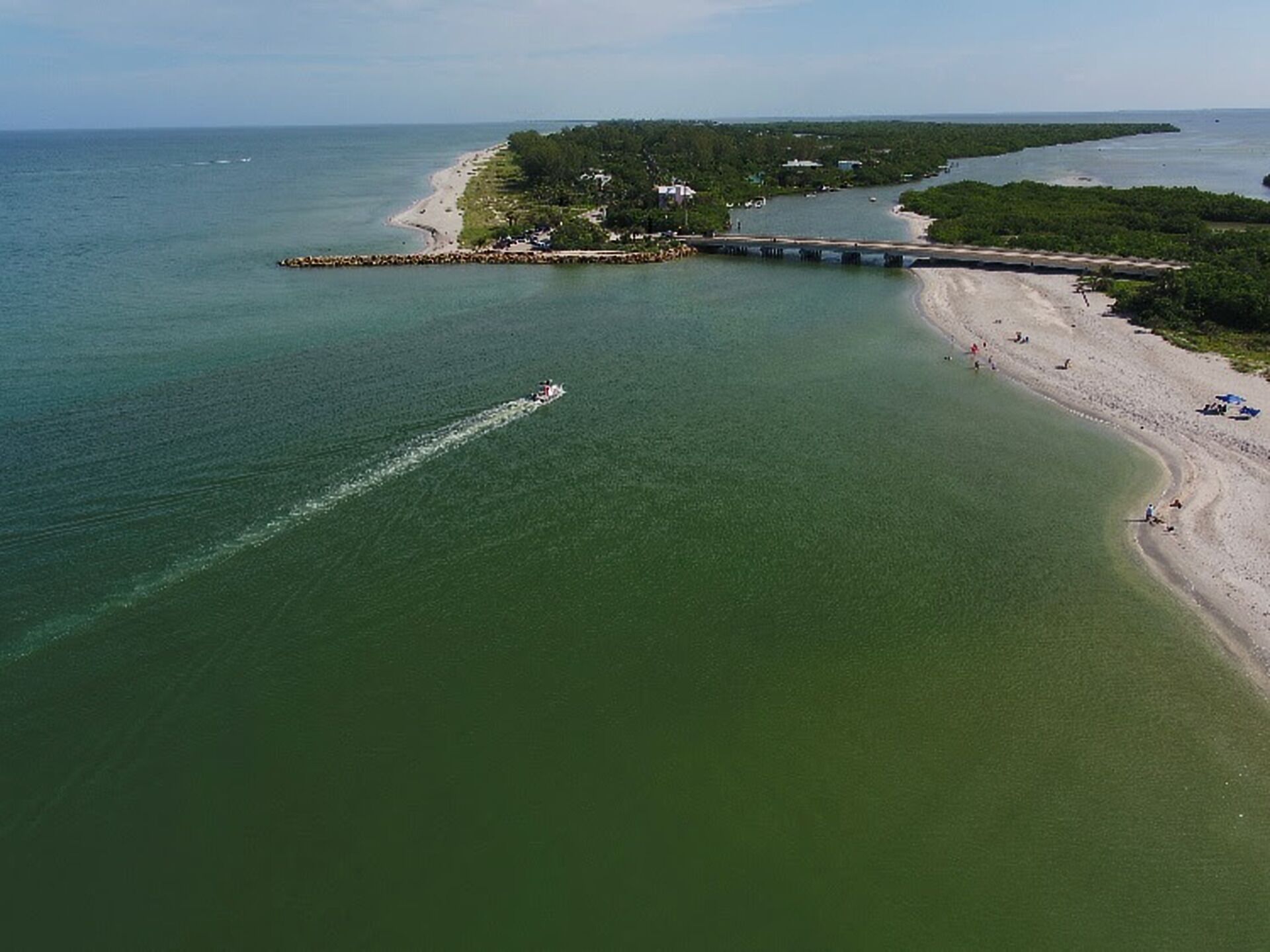 Ett flygbildsbild av ett kustområde med en strand, en pir och en båt som skapar en våg efter sig i vattnet.