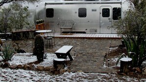 A stationary RV with a covered patio area, including a table and chairs, surrounded by cacti and rocks. - Unique Retreat - Airstream in Central Location - Remote Feeling. (Tucson)