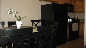 Eine kleine Küche mit Holzausstattung, einem schwarzen Kühlschrank und einer Mikrowelle. Ein Esstisch mit Stühlen und eine Orchidee im Blumentopf sind ebenfalls vorhanden.