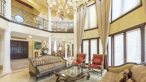 Amplia sala de estar elegantemente amoblada, con techo alto, candelabro ornamentado y grandes ventanales con cortinas.