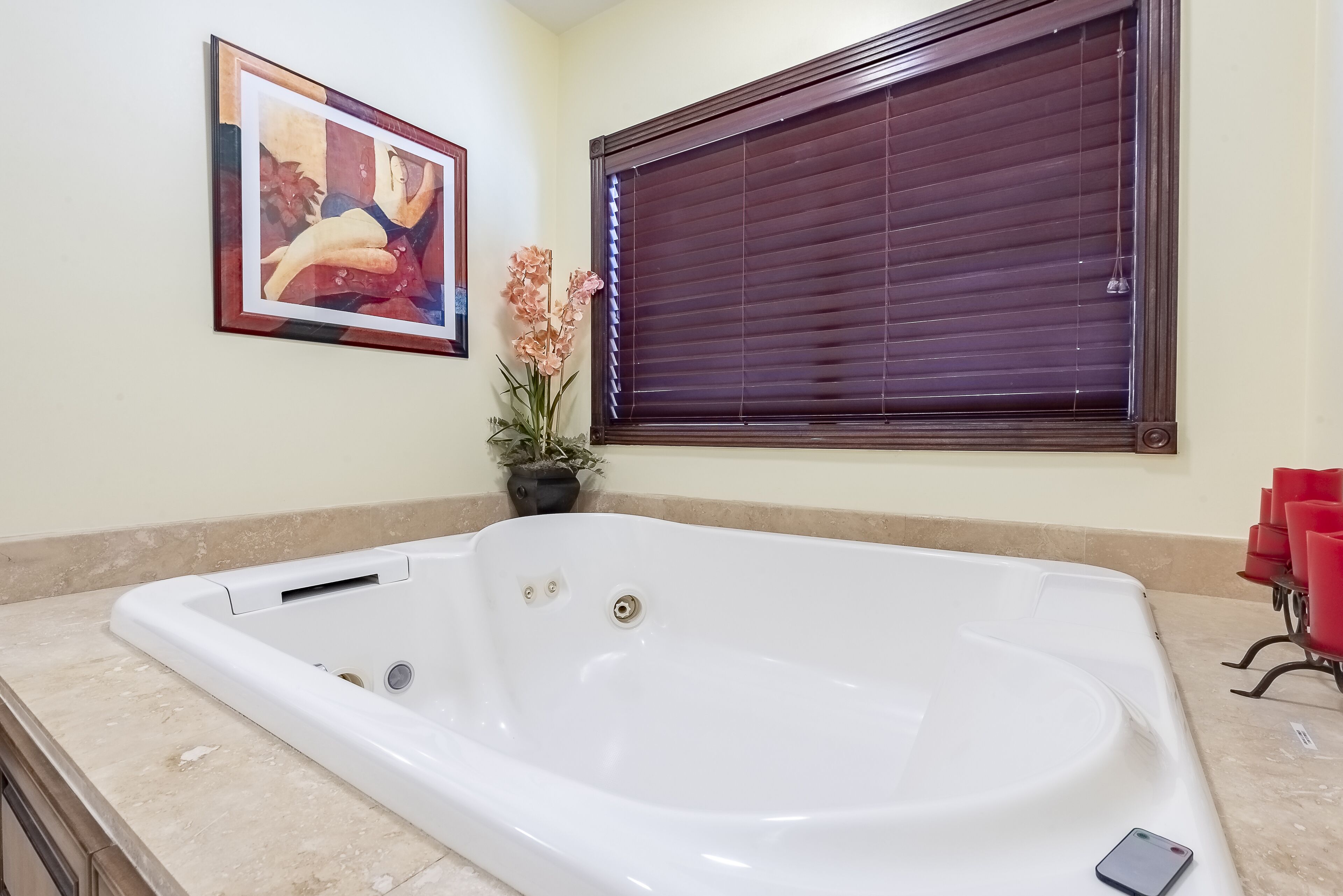A bathroom with a white bathtub, a framed painting, a potted plant, and a window with blinds.