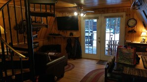 A wooden interior with a staircase, a flat-screen TV mounted on the wall, a door leading outside, and a clock on the wall. - Huckleberry Hideaway: Authentic Cabin Adventure, steps away from Payette Lake (McCall)