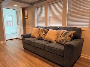 A grey sofa with patterned cushions, wooden flooring, a door, and windows with blinds.