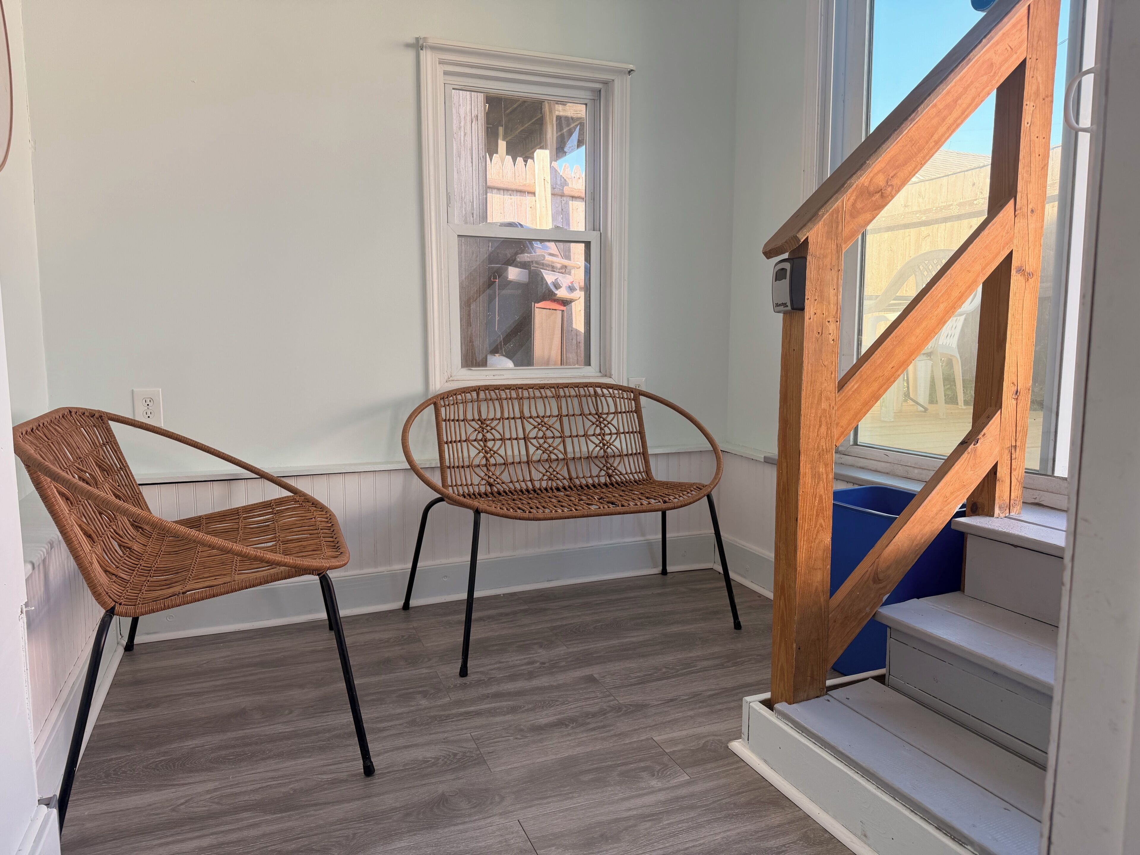 Una habitación con una escalera de madera, dos sillas de mimbre, un ventanal y una pared blanca.