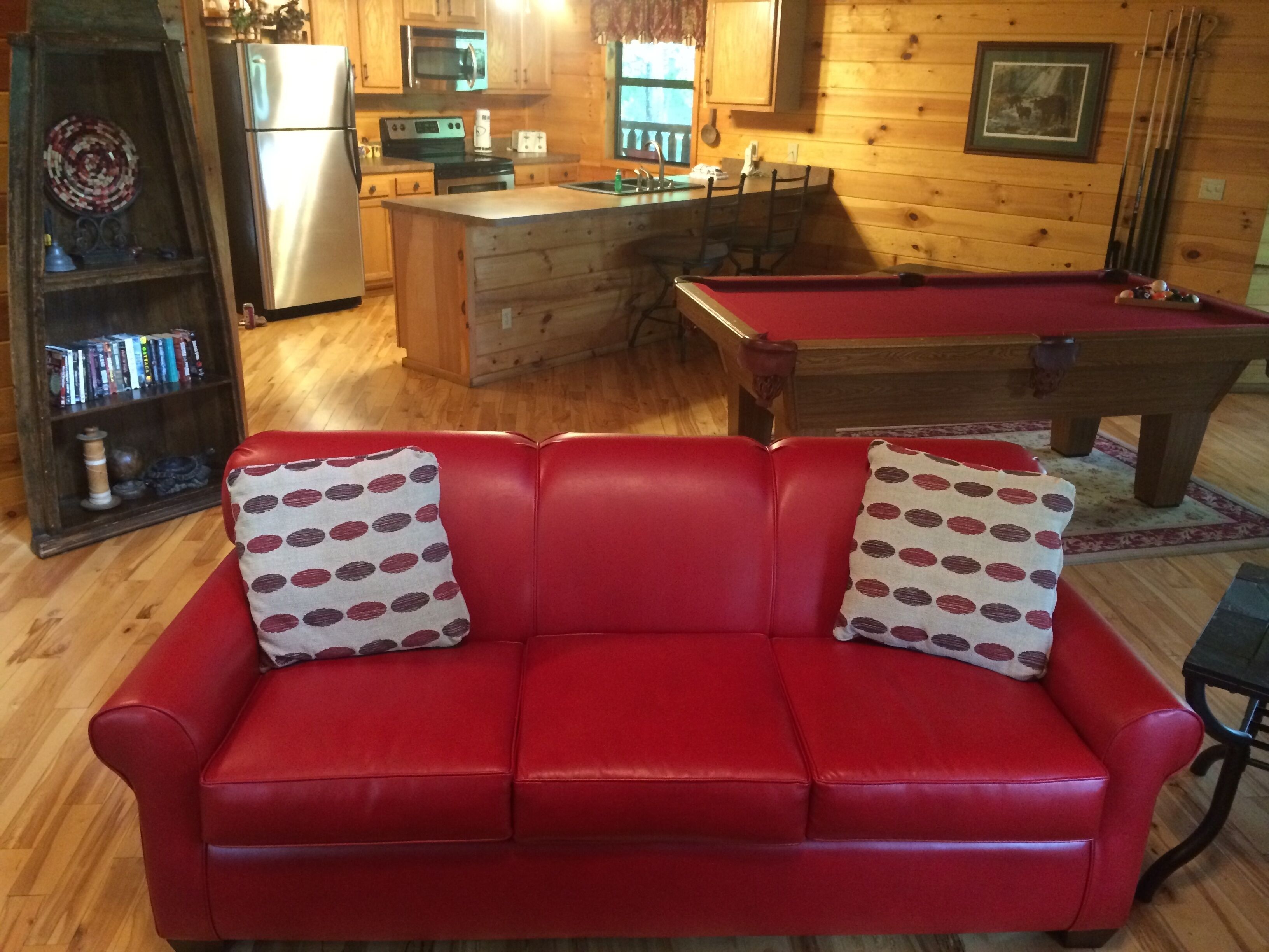 Una sala de estar acogedora con un sofá de cuero rojo, una mesa de billar, una estantería y una zona de cocina con armarios de madera y un refrigerador.