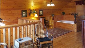 A cozy cabin interior with wooden walls, a wooden ceiling, a dining area with a table and chairs, a fireplace, and a large window.