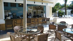 Un espace bar extérieur avec des meubles en osier et des tables en verre, une cuisine visible à travers une porte ouverte, et un coin salon au bord de la piscine avec des chaises longues.