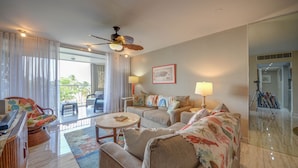 A spacious living room with a sofa, armchairs, a coffee table, and a ceiling fan. A balcony with curtains is visible through the glass doors.