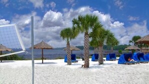 Een strand met een volleybalnet, palmbomen en ligstoelen onder rieten parasols.