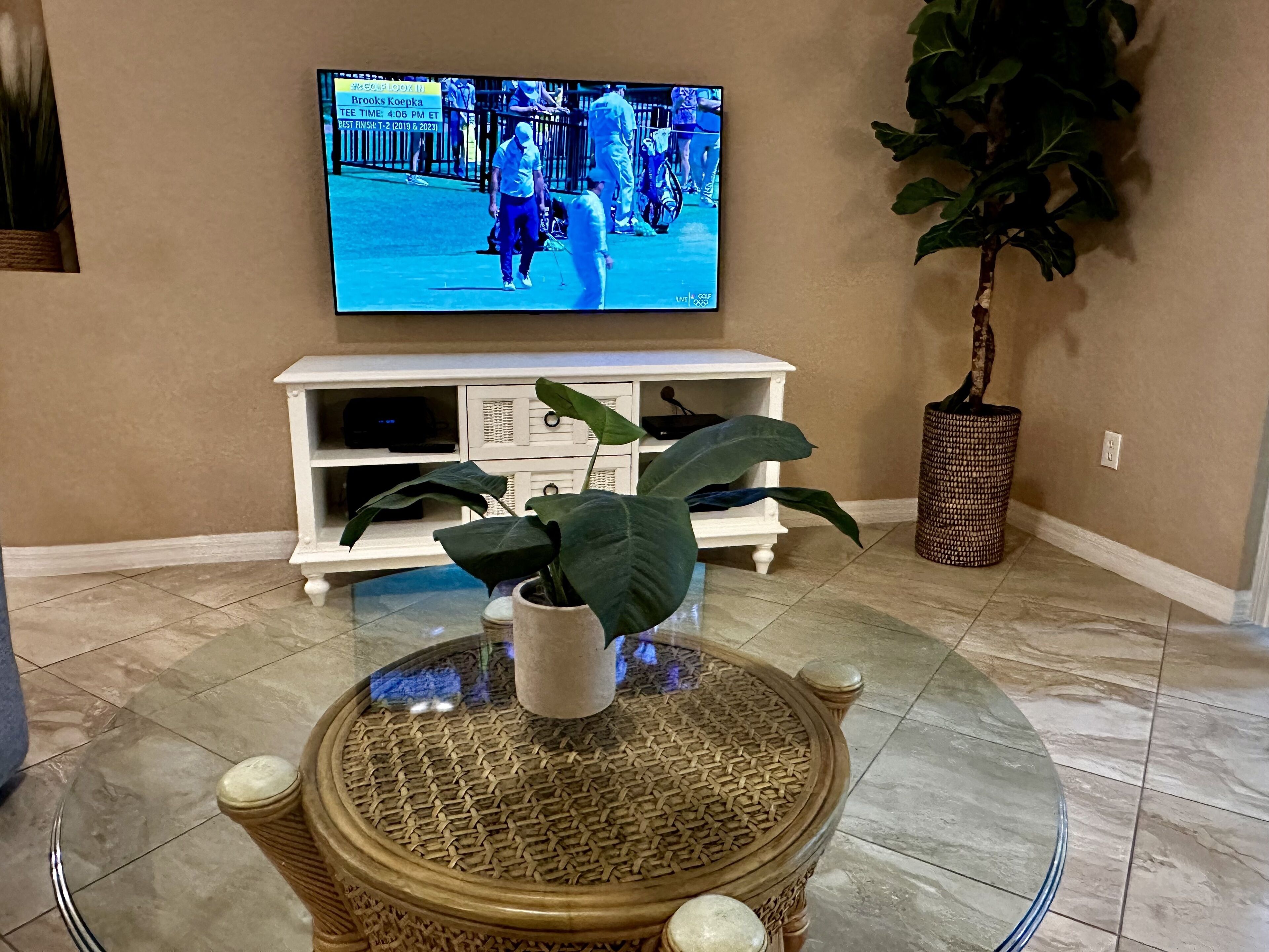 Un salon avec un téléviseur diffusant un événement sportif, un meuble de rangement blanc, une table basse ronde en osier, une plante en pot et un sol carrelé.