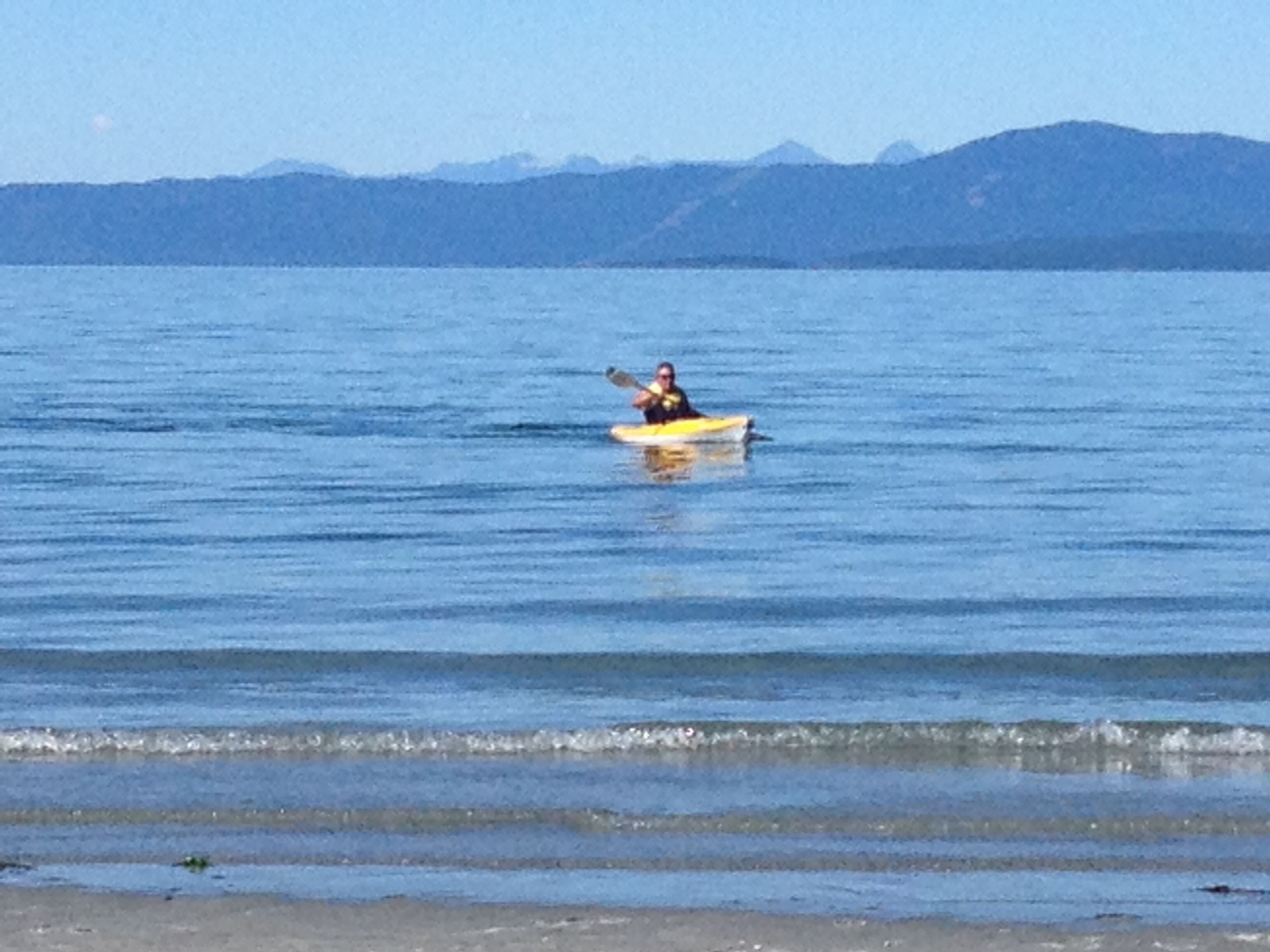 A person kayaking on calm water with mountains in the background.