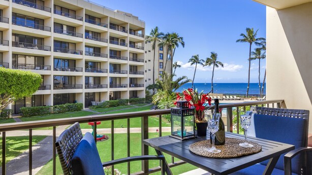 A balcony with a table set for two, overlooking a hotel building and the ocean. - SUGAR BEACH RESORT 10% DSCNT on 7 days Stay - Renovated Condo 201 (Kihei)