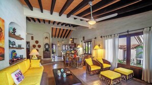 A spacious living area with a yellow sofa, wooden coffee table, and matching ottomans. A dining area with a wooden table and chairs is visible in the background. - Casa Colibri - Los Delfines (Sayulita)
