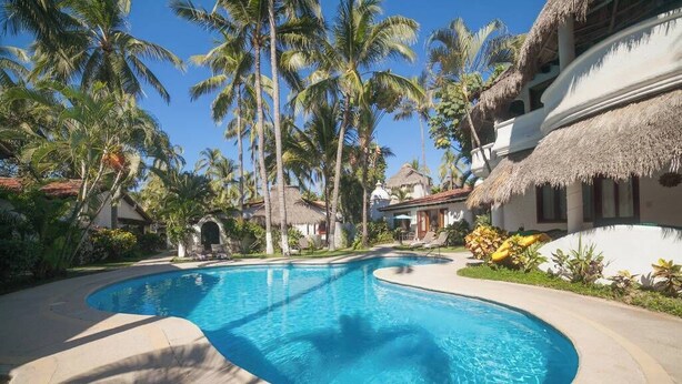A swimming pool with a curved edge, surrounded by palm trees and thatched-roof buildings. - Casa Colibri - Los Delfines (Sayulita)