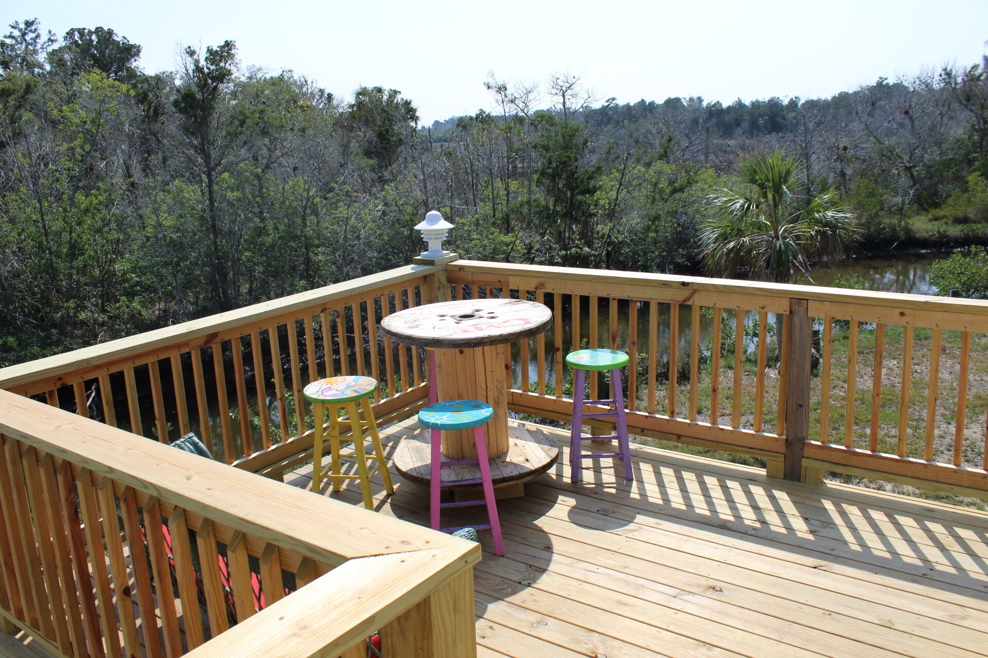 Eine Holzterrasse mit einem runden Tisch und drei bunten Hockern, mit Blick auf eine bewaldete Gegend und ein Gewässer.