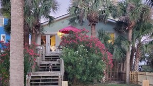 A wooden staircase leading to a house with a pink umbrella, surrounded by palm trees and flowering plants. - Beach is 50 steps away and you have PRIVATE beach access and wonderful views (Folly Beach)