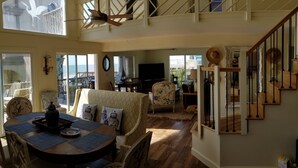 A spacious living area with a high ceiling, a dining table set with blue placemats, and a view of the ocean through large windows. - Beach is 50 steps away and you have PRIVATE beach access and wonderful views (Folly Beach)
