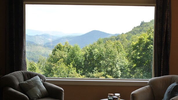 A room with a large window offering a view of a forested mountain range. There are two chairs and a small table with jars on it.