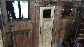 A wooden cabin interior with a wooden door, a window, a shelf with a candle, and a metal ladder.